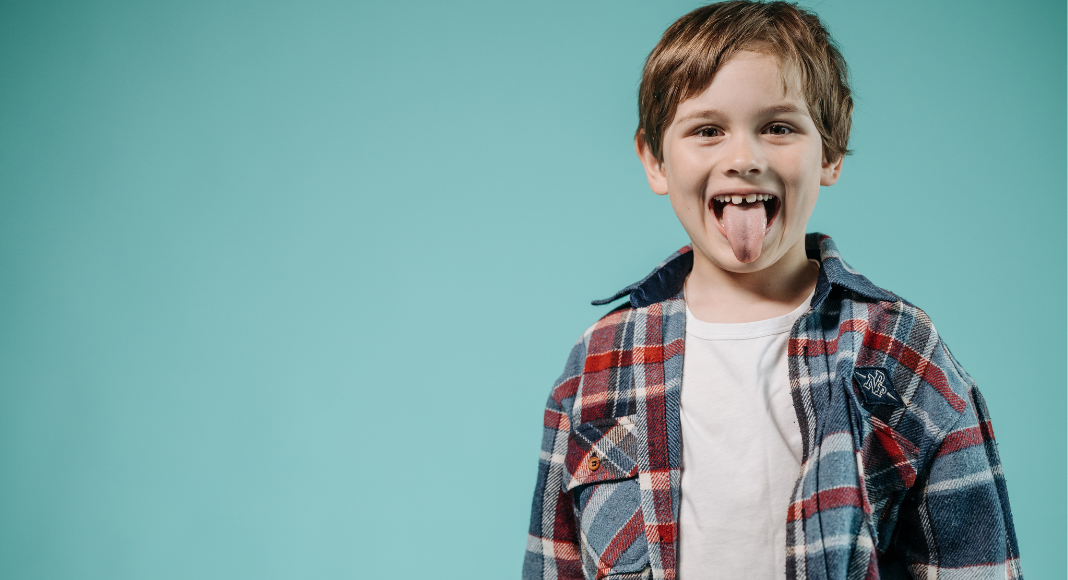 Boy with tongue out wearing a plaid shirt against a teal background. parenting lessons. Des Moines Mom